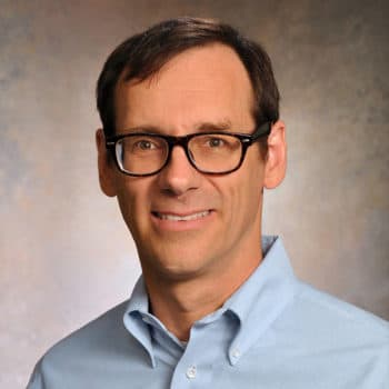 Smiling man with short dark hair, glasses, and a light blue collared shirt.