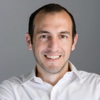 Smiling man with short dark hair in a white shirt against a gray background.