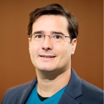 Smiling man with dark hair, glasses, wearing a blue jacket and teal sweater.