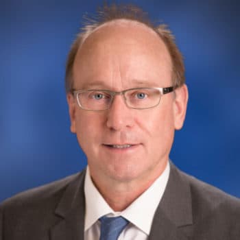 Smiling man with glasses, gray suit, and blue tie against a blue background.